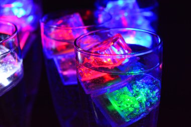 Farklı renklerde parlayan buz küpleri olan uzun burkulmuş bacaklarda beş cam kadeh. Aydınlık elementlere sahip eğlenceli ve parlak içecekler siyah bir arka plan, parti ve rahatlama üzerinde yer alır..