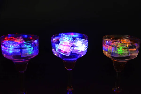 Three open champagne glasses with luminous multicolor ice cubes are located on a black matte background.