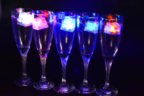 Five glass goblets on a long twisted legs with ice cubes glowing in different colors. Fun and bright drinks with luminous led elements are located on a black matte background, party and relaxation.