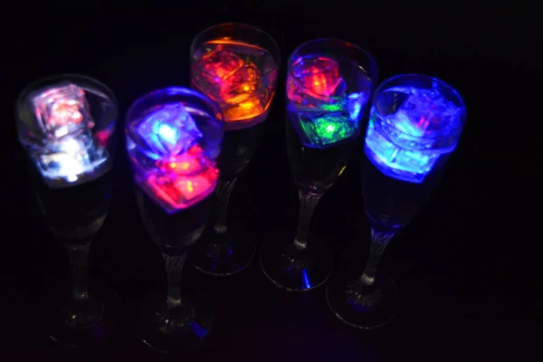 Five glass goblets on a long twisted legs with ice cubes glowing in different colors. Fun and bright drinks with luminous led elements are located on a black matte background, party and relaxation.