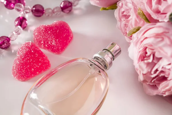 Bottle of perfume, flowers and hearts on a light background. Close-up bottle of perfume on the background of flowers and two pink sweet candies in the shape of a heart. Pastel colors, a gift for Valentine\'s Day.