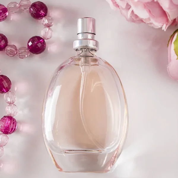Perfume bottle and flowers on a white background. A beautiful bottle of perfume on a light background surrounded by beads and rose flowers. Square photo, pastel colors.