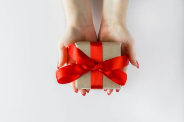 Beyaz arka planda kırmızı kurdeleli bir hediye. Kadınların elinde Sevgililer Günü hediyesi. El işi kağıdında hediye kutusu ve kırmızı saten kurdele..