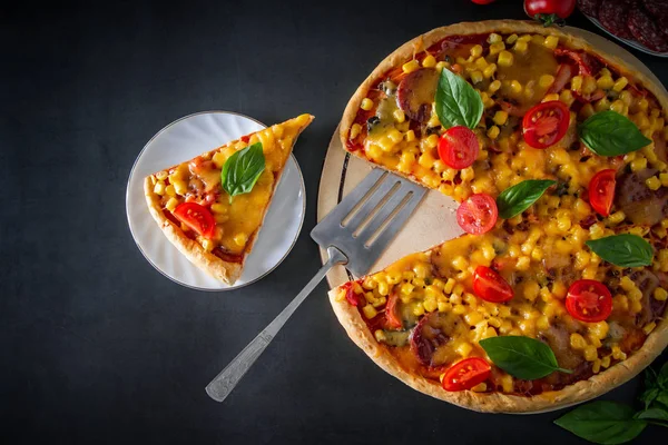 Slice of pizza with tomatoes and basil on a black background. National Italian food, pizza with tomatoes, corn, green basil and cheese on a round board. Vertical photo, place for text.