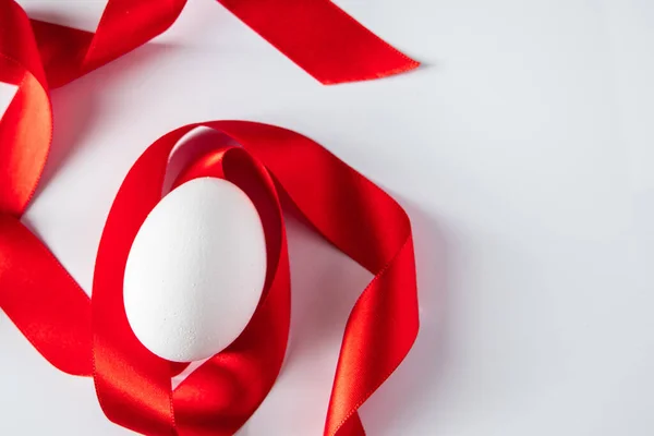 White egg with red silk ribbon. Easter symbol. Closeup white egg with red silk ribbon, easter symbol on a light background. Place for text.