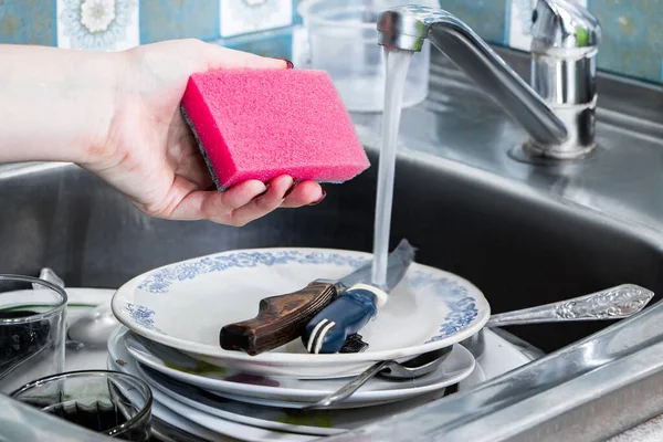 A young woman washes dirty dishes and holds in her hand a pink sponge with foam. Homework cleaning.