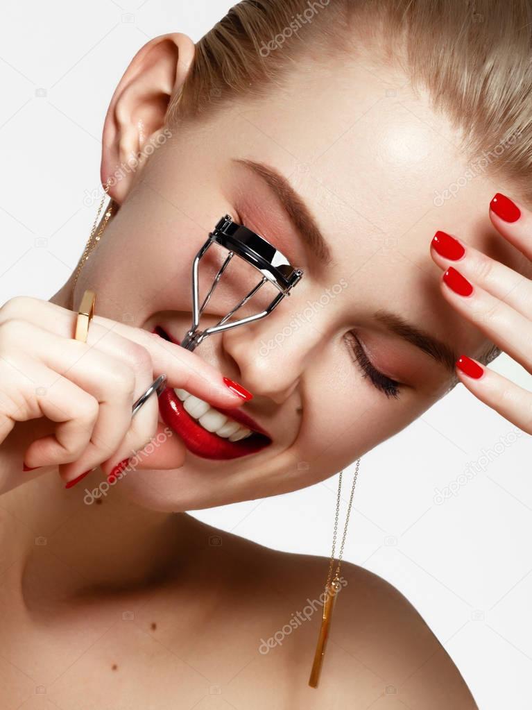 Makeup products. Young beautiful girl with gold earrings and ring smiling on white background. Red nails with manicure and white smile. A young woman with fashion accessories applying eyelash curler