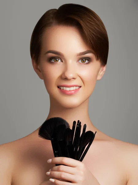 Krásná mladá žena použití nadace na její tváři s make-up štětce izolované na šedém pozadí — Stock fotografie