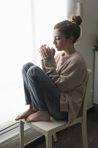 Chica adolescente sentada junto a una ventana con una taza de café en la mano . — Foto de Stock