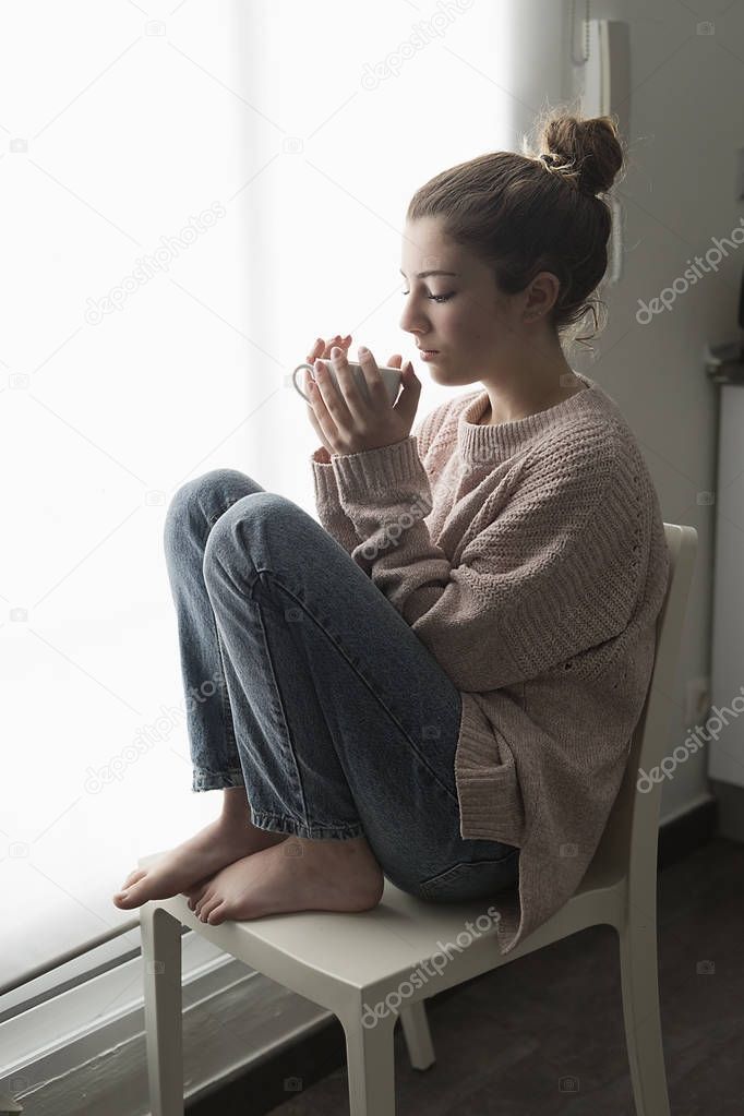Teen girl sitting by a window with a cup of coffee in her hand.