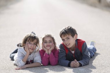 Kamera karşı karşıya bir yol yerde yatan üç çocuk.