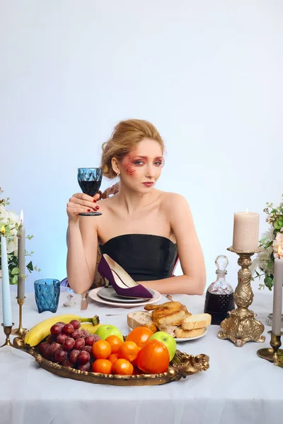 Modelo loira de moda com maquiagem colorida bebendo vinho atrás da mesa decorada e comendo sapato de camurça — Fotografia de Stock