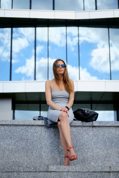 Young attractive stylish brunette woman sitting on marble surface. Reflection of the sky in windows. — Stock Photo, Image