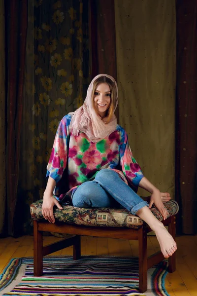 Carino signora in sciarpa di seta intorno alla testa in posa sul divano con tessuto colorato sullo sfondo — Foto Stock