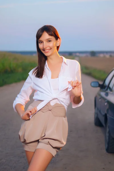 Chica atractiva en camisa blanca posando cerca de coche en la carretera del campo en la puesta del sol — Foto de Stock