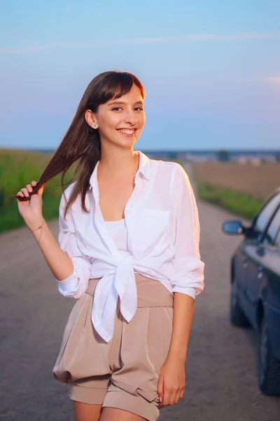 Menina atraente na camisa branca posando perto do carro na estrada do país ao pôr do sol — Fotografia de Stock