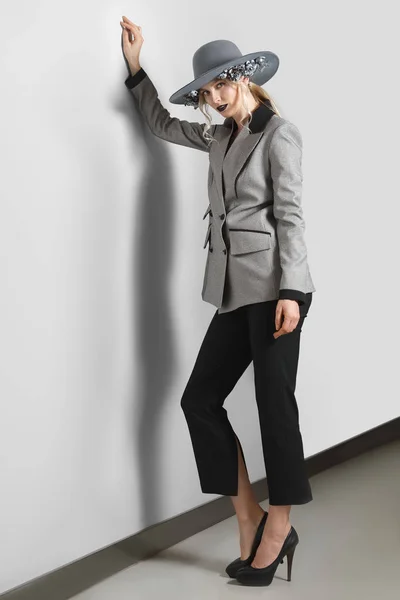 Bonita modelo de moda en chaqueta, pantalones y sombrero de ala ancha con flores posando cerca de la pared gris — Foto de Stock