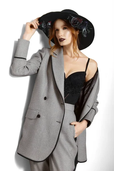 Hübsches Mode-Model in Jacke, Hose und breitkrempigem Hut mit Blumen, die in der Nähe der grauen Wand posieren — Stockfoto