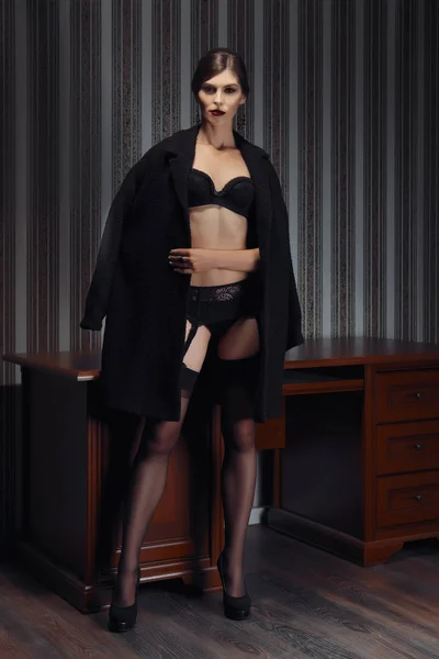 Hermosa dama en lencería negra y medias con cinturón de tirante y abrigo de pie en habitación oscura junto a la mesa —  Fotos de Stock