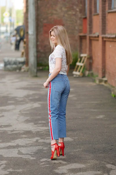 Привлекательная блондинка в коротких джинсах, кружевной блузке и красных туфлях позирует на улице — стоковое фото