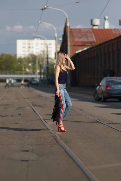 Søt, blond jente med solbriller som krysser veien med skinnjakke i hånden. – stockfoto
