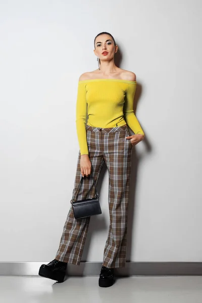 Fille en pantalon à carreaux et pull jaune avec petit sac à main posant près du mur blanc — Photo