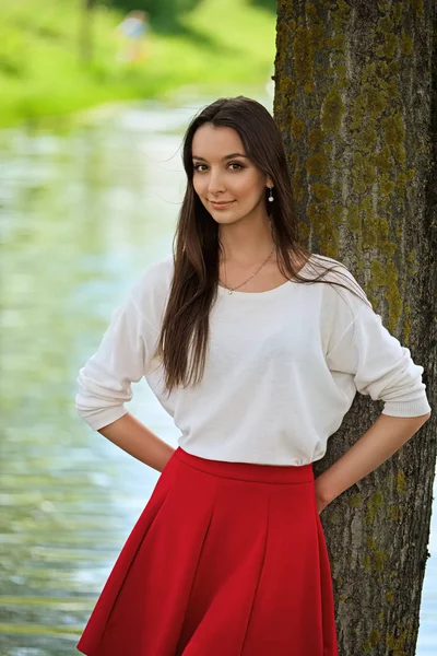 Πορτρέτο ενός κοριτσιού με κόκκινη φούστα και άσπρη μπλούζα ακουμπισμένη σε ένα δέντρο στην όχθη του ποταμού — Φωτογραφία Αρχείου