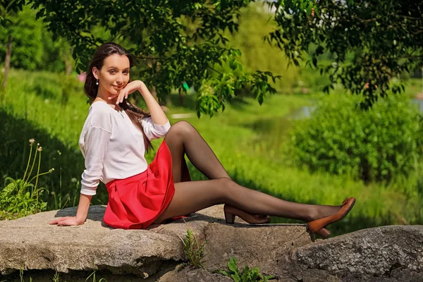 Retrato de una chica en falda roja y blusa blanca sentada en piedra en la orilla del río — Foto de Stock