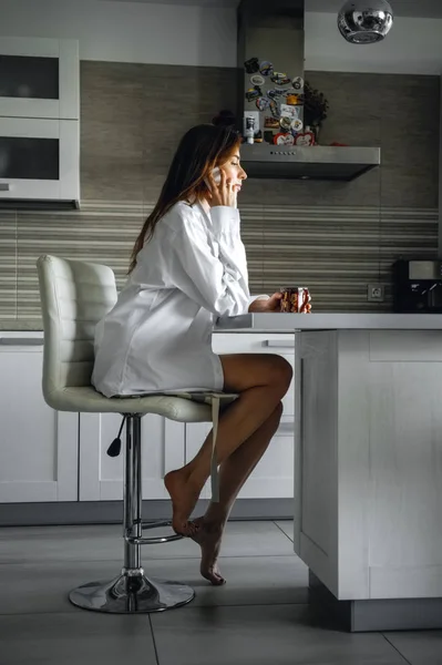 Beautiful girl in white shirt sitting with cup of tea in the kitchen and talking on mobile phone — 스톡 사진