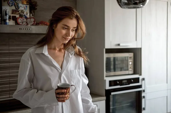 Gyönyörű lány fehér ing ül egy csésze tea a konyhában, és beszél a mobiltelefon — Stock Fotó