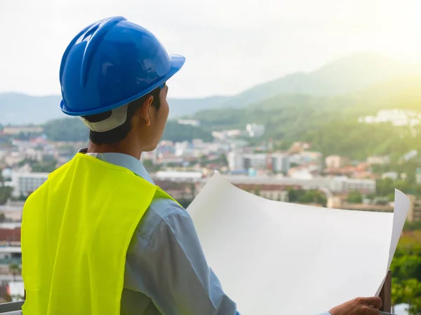 A construction engineer. Wearing a helmet and vest holding a plans with blur city or building background in construction concept