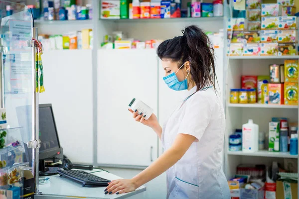 the pharmacist is holding a container with medicine and is studying it. A pharmacist in a medical mask on his face punches medicine in a computer.