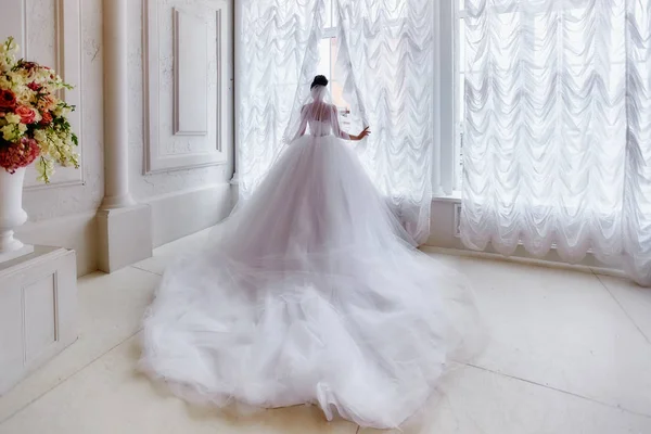 Die Braut steht am Fenster in einem Kleid mit langer Schleppe und — Stockfoto