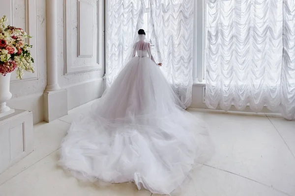 Beautiful bride with a long train, standing at the window. — Stock Photo, Image