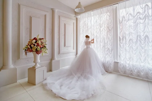 Schöne Braut mit langer Schleppe, die am Fenster steht. — Stockfoto