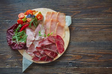 Çeşitli et lezzetleri, yuvarlak bir tahta üzerinde ince dilimler halinde kesilmiş. Sebze ve taze otlarla süslenmiş bruschetta.