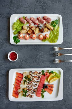 Çeşitli balıklar, kırmızı balık kanepeleri, patlıcan ve jambonlu börekler. Gri arka planda beyaz bir tabakta peynir ve otlarla doldurulmuş..