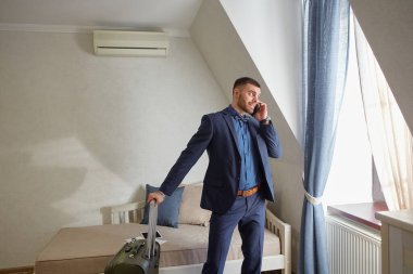 Resmi giyinmiş genç bir işadamı pencerenin yanında telefonla konuşuyor. Bavul toplandı. Gitmeye ve yeni bir iş gezisine başlamaya hazırım..