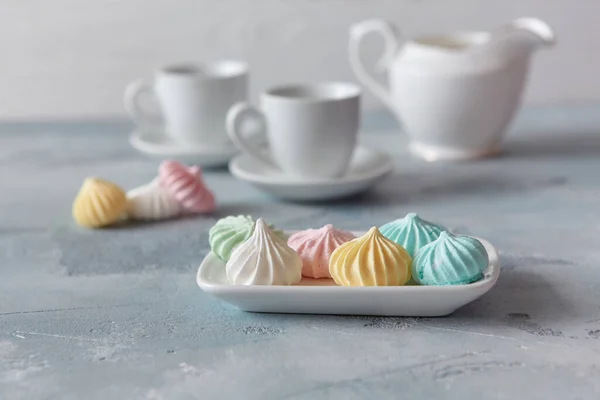 Multi-colored meringues in pastel shades with cups and a jug on a light textured wooden background. The concept of Breakfast with tea or coffee, home comfort and tenderness. Close-up, selective focus.