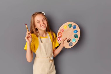Güzel bir genç kızın portresi, tahta bir palet tutuyor ve gri bir duvara sürtünüyor. Elinde sanat paleti ve fırçası olan gülümseyen bir çocuk. Çocukların yaratıcılığı için eşyalar..