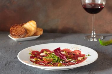 Ahşap zemin üzerinde beyaz bir tabakta et carpaccio, lezzetli bir ekmek ve bir bardak şarap ile.