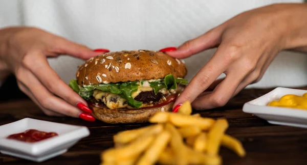 Hands holding fresh delicious burgers with french fries and sauce on the wooden table. Homemade burgers in a rustic style. Cheeseburger, with beef, tomato, cheese, cucumber and lettuce. Soft focus.