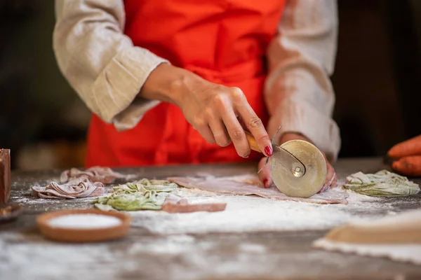 Senior woman cook making tagliatelle pasta with dough in restaurant kitchen. Making pasta. Making pasta in restaurant. Colored raw vegetable pasta with beets, carrots and spinach.