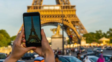 Paris 'teki Eyfel Kulesi. Güneş ışığı ve mavi gökyüzü altında. Dünyaca ünlü popüler turist mekanı. Paris 'te turist Fransa' yı geziyor, kadın cep telefonuyla fotoğraf çekiyor..