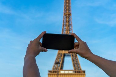 Paris 'teki Eyfel Kulesi. Güneş ışığı ve mavi gökyüzü altında. Dünyaca ünlü popüler turist mekanı. Paris 'te turist Fransa' yı geziyor, kadın cep telefonuyla fotoğraf çekiyor..