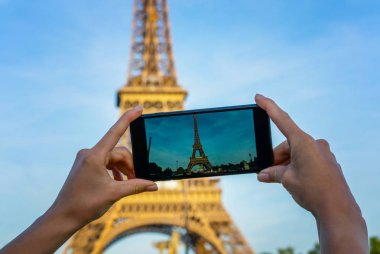 Paris 'teki Eyfel Kulesi. Güneş ışığı ve mavi gökyüzü altında. Dünyaca ünlü popüler turist mekanı. Paris 'te turist Fransa' yı geziyor, kadın cep telefonuyla fotoğraf çekiyor..