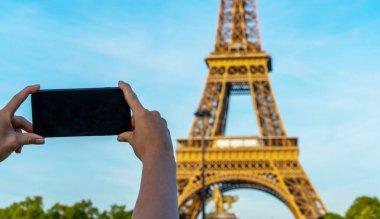 Paris 'teki Eyfel Kulesi. Güneş ışığı ve mavi gökyüzü altında. Dünyaca ünlü popüler turist mekanı. Paris 'te turist Fransa' yı geziyor, kadın cep telefonuyla fotoğraf çekiyor..