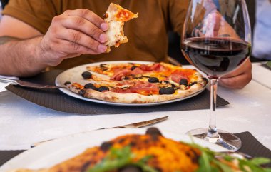 Tabakta geleneksel İtalyan pizzası var. Restoranda pizza yiyen bir adam. Adamın elleri taze pizzayı domates ve zeytinle alıyor..