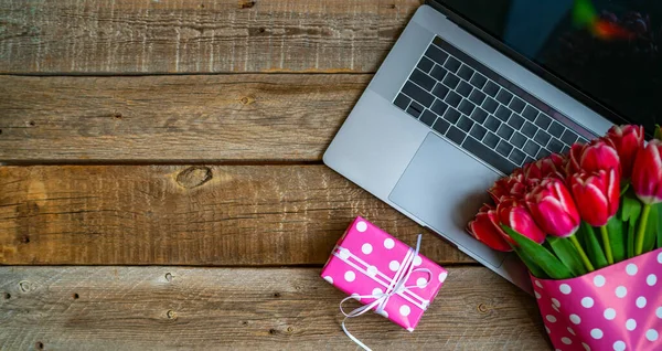 Working space or working place with laptop, flowers and gift box on rustik wooden background with copy space. Spring or business concept. Top view, flat lay.