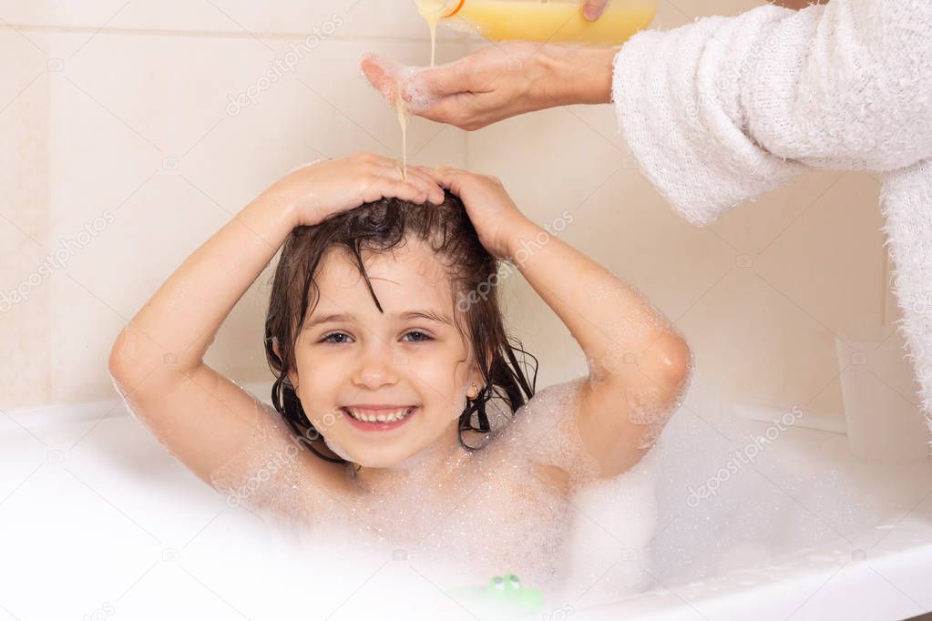 Shampoo hair and soap for children. Kid bathing in large tub. Baby girl with foam in hair. Mother washing child hair with shampoo in the shower.